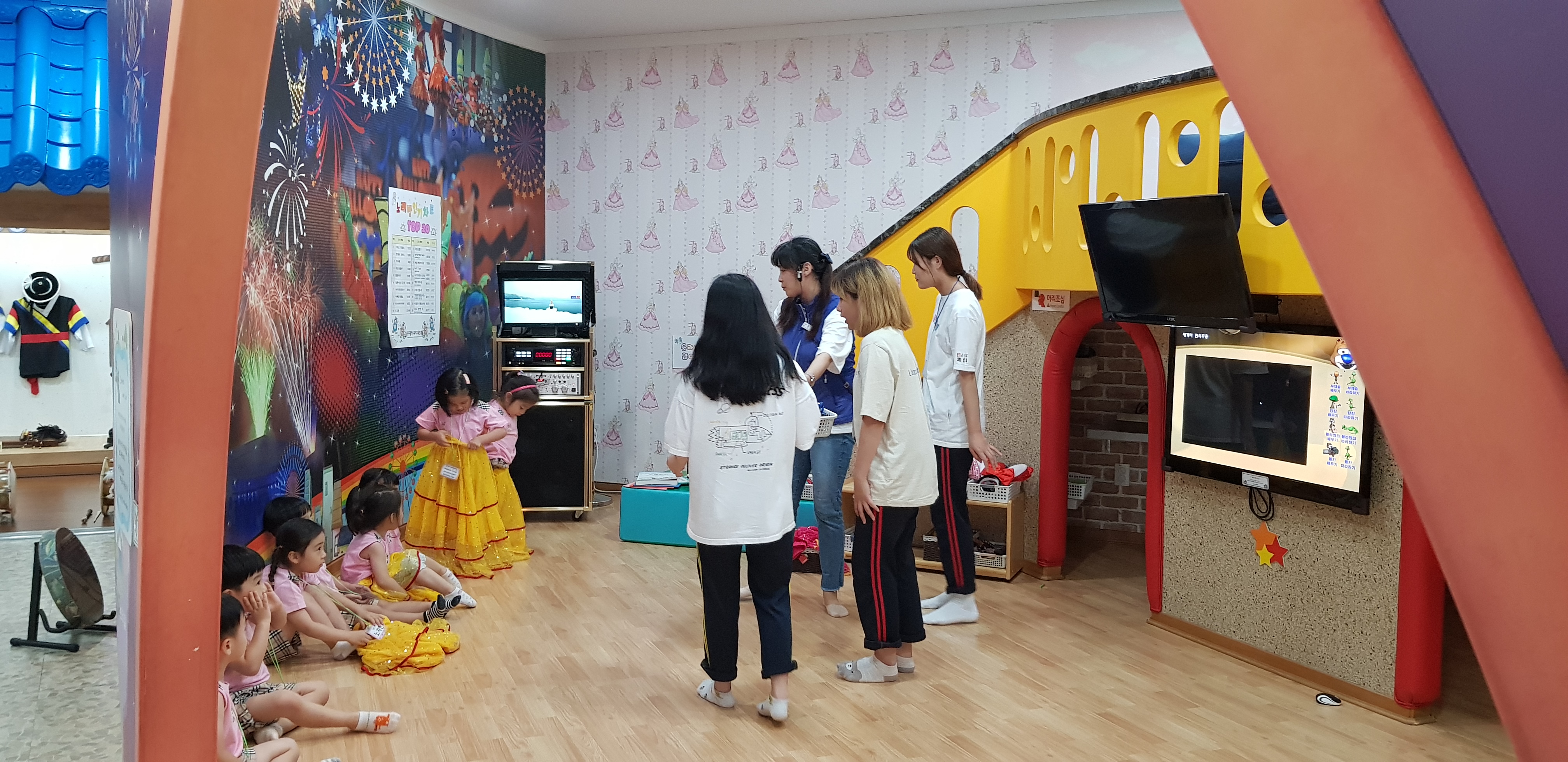 체험의상을 입고있는 유아들과 앉아있는 유아들 과 설명하는 선생님 옆에 서있는 고등학생들