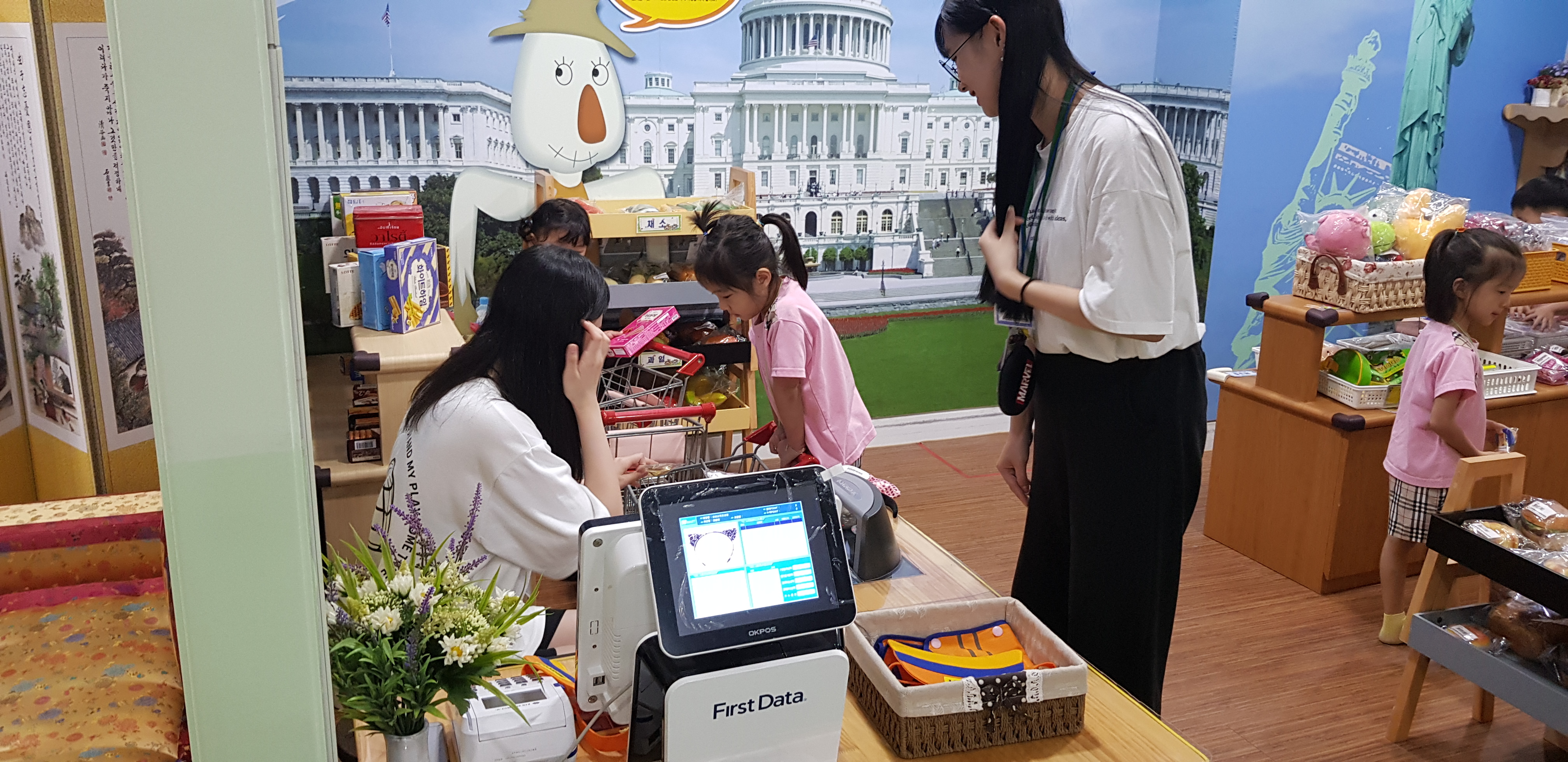 계산대쪽에서 유아들을 보고있는 고등학생 두명과 체험하고있는 유아들