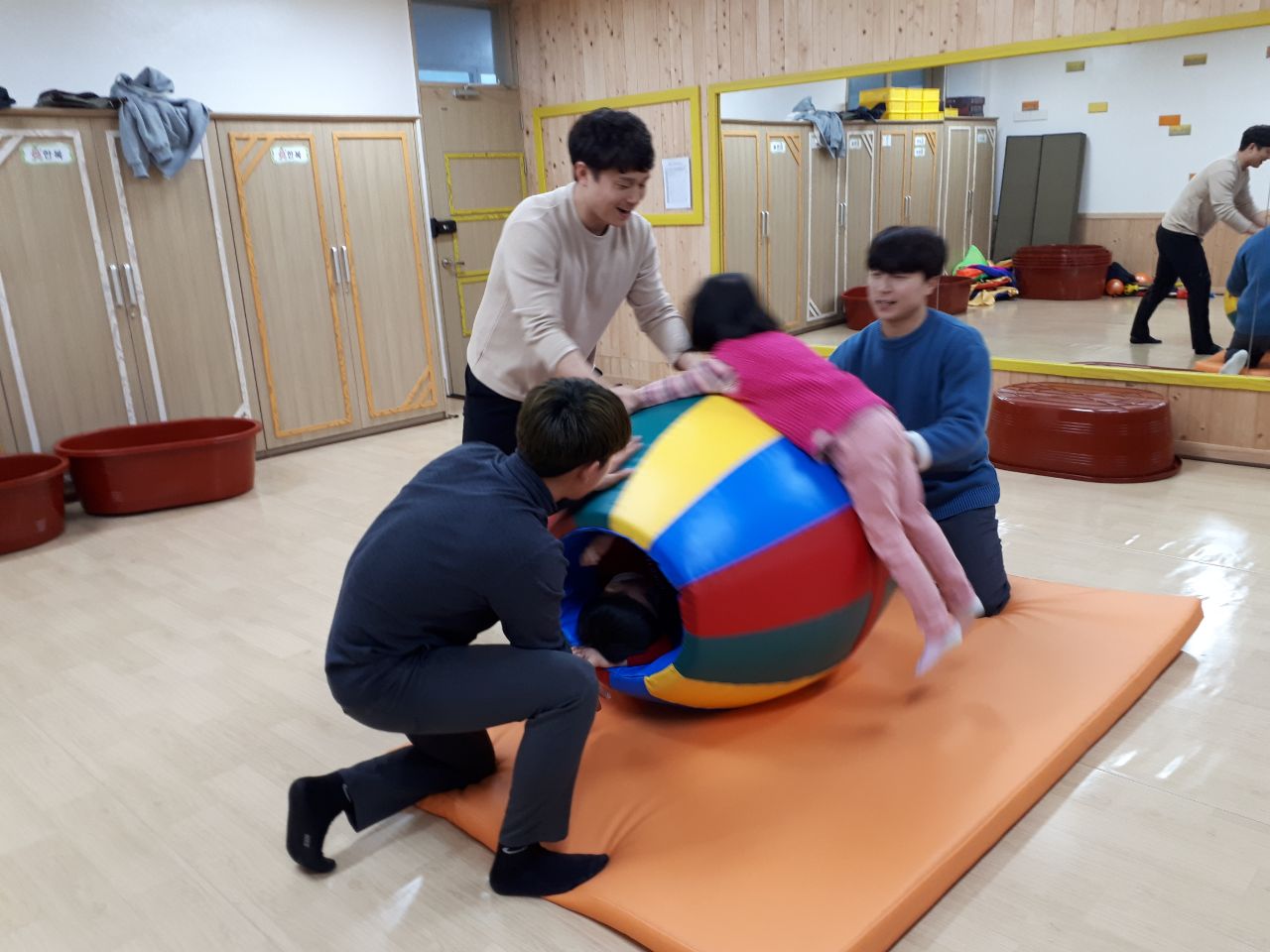 원형 놀이기구를 눕혀서 그위에 올라탄 여자아이와 그걸 도와주는 세 남성